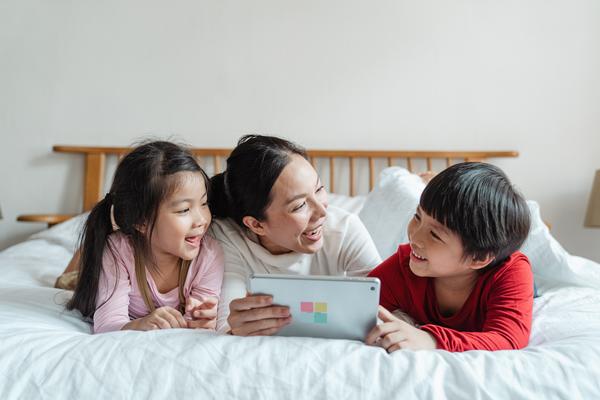 Mother and two children lying on white bed looking at electronic tablet