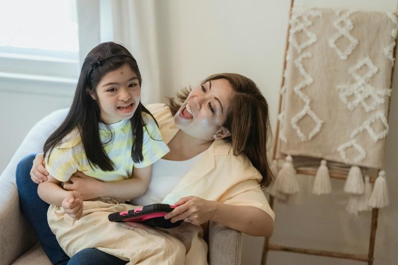 A child sits on a parent's lap and they both look excited in conversation, the parent holding a tablet